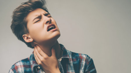A young man in a plaid shirt expresses visible discomfort as he holds his throat, showcasing a relatable moment of health distress or anxiety in a minimalistic setting.の素材