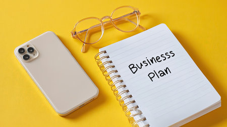 A modern workspace featuring a notebook titled "Business plan," stylish glasses, and a smartphone on a vibrant yellow background, inspiring creativity and productivity.の素材