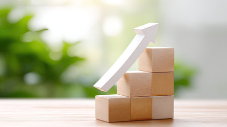 A creative representation of business growth featuring wooden blocks stacked in steps with a white upward arrow against a blurred green background symbolizing success and progress.の素材