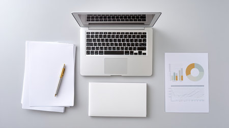 A clean and minimalist workspace featuring a laptop, blank papers, a pen, and a graphical report on a grey background, perfect for business planning.の素材