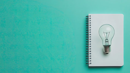 A light bulb rests on an open notebook against a teal background, symbolizing creativity and innovation. Perfect for educational or business themes.の素材