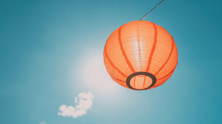 A bright orange paper lantern hangs gracefully against a clear blue sky, radiating warmth and charm. This vibrant decoration symbolizes celebration and culture.の素材