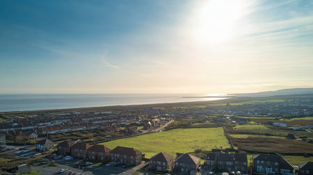 Captivating aerial view of a coastal landscape at sunset, showcasing calm waters reflecting the sun. A peaceful scene with rural fields and residential buildings.の素材