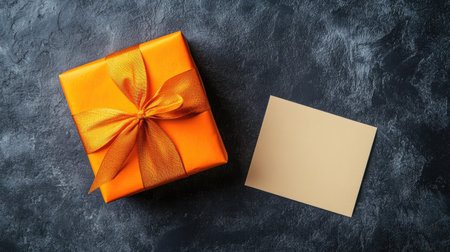 A vibrant orange gift box with a textured ribbon sits next to a blank card on a dark surface, perfect for any celebration or special occasion.の素材