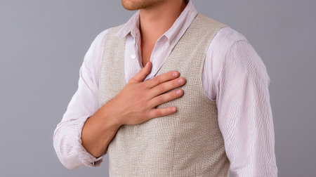 A man wearing a light-colored vest stands against a neutral backdrop, placing his hand on his chest, signifying sincerity and emotion.の素材
