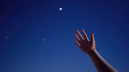 A hand stretches towards the night sky, filled with stars and a glowing moon, capturing a moment of aspiration and wonder against a deep blue backdrop.の素材