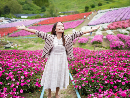 Joyful woman stands in vibrant flower field, surrounded by colorful blooms. She wears light dress and striped cardigan, with arms outstretched, embracing beauty of natureの写真素材