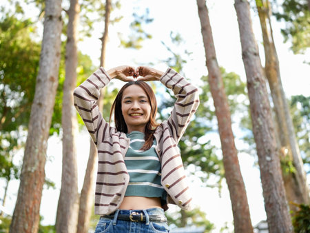 Young woman stands outdoors among tall trees, smiling and forming heart shape with her hands. scene conveys joy and connection with nature, highlighting carefree momentの写真素材