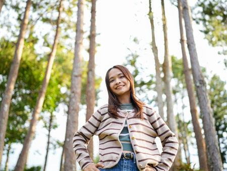 Young woman stands confidently in forest, surrounded by tall trees and greenery. She wears striped cardigan and smiles warmly, embodying sense of joy and connection with natureの写真素材