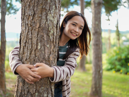 Young woman smiles while embracing tree in serene forest setting. sunlight filters through trees, creating warm and inviting atmosphereの写真素材