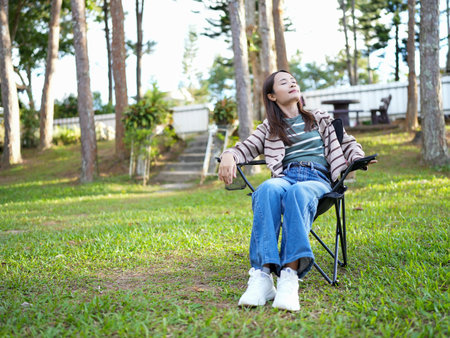 Relaxing in chair, woman enjoys serene outdoor setting surrounded by tall trees and greenery. peaceful atmosphere invites tranquility and reflectionの写真素材