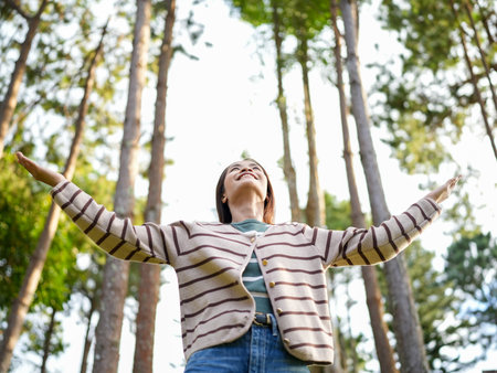 Joyful woman stands in forest, arms outstretched, embracing nature. Sunlight filters through tall trees, creating serene atmosphere. Her expression reflects happiness and freedomの写真素材