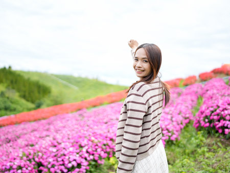 Young woman stands in vibrant flower field, smiling and waving. landscape features colorful blooms in shades of pink, creating cheerful and lively atmosphereの写真素材