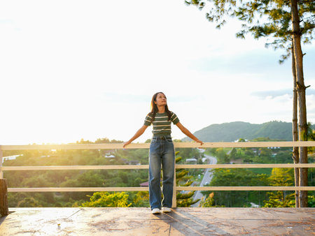 Young woman stands on balcony, enjoying scenic view of rolling hills and sunset. warm light creates serene atmosphere, highlighting her relaxed expression and casual outfitの写真素材