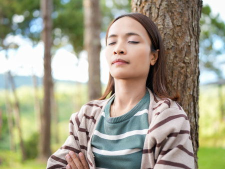 Woman is meditating peacefully against tree serene outdoor setting. She has her eyes closed, exuding calmness and tranquility. lush greenery background enhances peaceful atmosphere, making itの写真素材