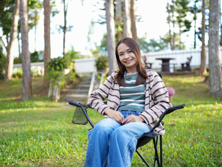 Young woman sits comfortably in folding chair outdoors, surrounded by trees and greenery. She wears striped sweater and jeans, exuding relaxed and cheerful vibeの写真素材
