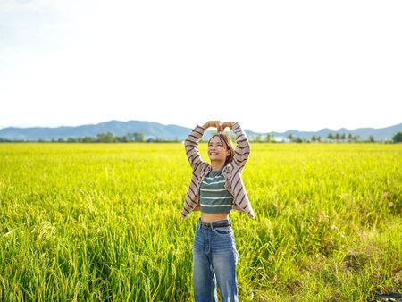 Young woman stands in lush green rice field, smiling and forming heart shape with her hands. bright sky and distant mountains create serene backdrop, evoking feelings of joy and freedomの写真素材