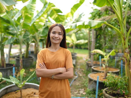 A confident young woman stands in a lush garden, surrounded by thriving banana plants. Her bright orange shirt contrasts beautifully with the vibrant greenery, reflecting a passion for nature.の写真素材