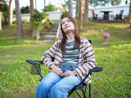 Young woman sits comfortably in folding chair, gazing thoughtfully upwards in serene outdoor setting. lush green grass and trees create peaceful atmosphere, perfect for relaxationの写真素材