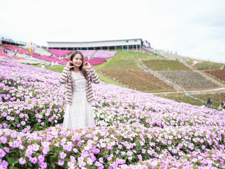 Young woman stands joyfully in vibrant flower field filled with pink blooms, surrounded by lush greenery and scenic hillside. atmosphere is cheerful and inviting, perfect for day outdoorsの写真素材