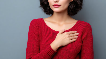A young woman in a cozy red sweater gently places her hand on her heart, radiating sincerity and warmth, perfect for themes of emotional connection and personal care.の素材