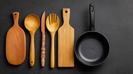 A visually appealing lineup of various wooden kitchen utensils, including spoons and cutting boards, alongside a sleek non-stick frying pan on a dark background.の素材
