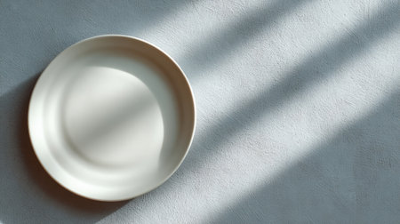 A minimalist white plate elegantly rests on a textured surface, illuminated by soft natural light, accentuating its simple beauty and inviting a serene culinary atmosphere.の素材