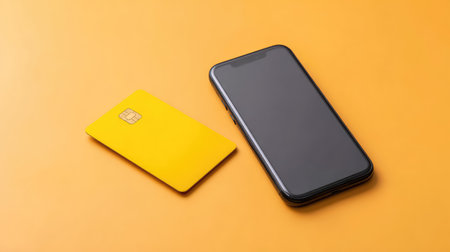 A vibrant yellow credit card and a sleek smartphone are placed on a bright orange background, symbolizing modern financial transactions and digital banking solutions.の素材