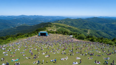 A breathtaking aerial view of a vibrant outdoor music festival set in rolling green mountains, filled with attendees enjoying the sunny day in a festive atmosphere.の素材