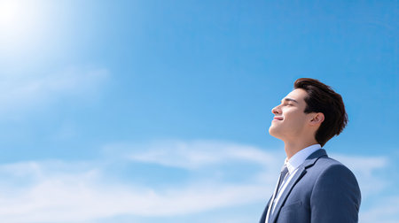 A young man in a blue suit stands outdoors under a clear sky, basking in sunlight with a serene smile, reflecting feelings of happiness and personal growth.の素材