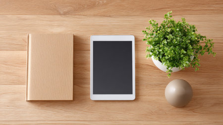 A minimalist workspace featuring a tablet, a book, a small green plant, and a decorative ball on a wooden table, perfect for creating a calm and organized environment.の素材