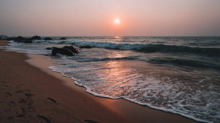 A mesmerizing sunset casts a warm glow over gentle waves lapping at a serene beach, where soft sand meets silhouetted rocks, creating a calming nature scene.の素材
