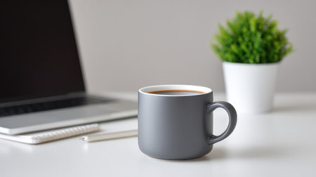 A stylish gray coffee cup sits on a bright desk beside a laptop and a small green plant, creating a perfect atmosphere for work or relaxation in a modern workspace.の素材