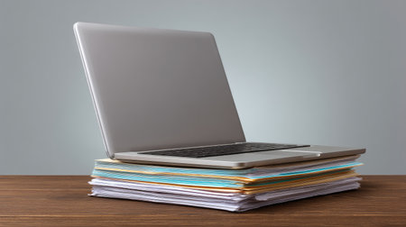 A modern silver laptop is positioned on an organized stack of colorful papers and folders, set against a neutral gray background, ideal for office or study settings.の素材