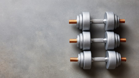 A clean arrangement of silver dumbbells on a smooth gray surface, ideal for fitness-related content, illustrating strength training and healthy lifestyle choices.の素材