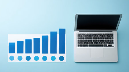 A clean and modern workspace featuring a laptop next to a blue business growth chart, symbolizing financial progress and productivity in a professional environment.の素材