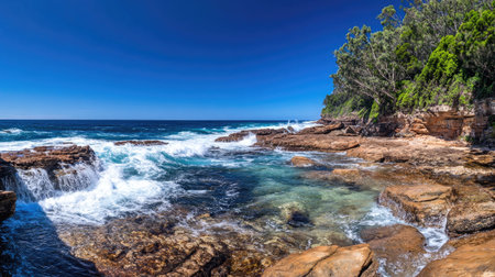 A panoramic view of a rugged coastline with waves crashing against the rocks and a clear blue sky.の素材