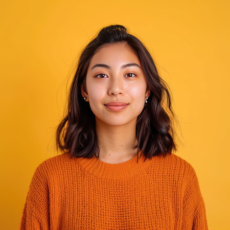 A young adult in a casual orange sweater with a neutral expression stands against a bright yellow backgroundの素材