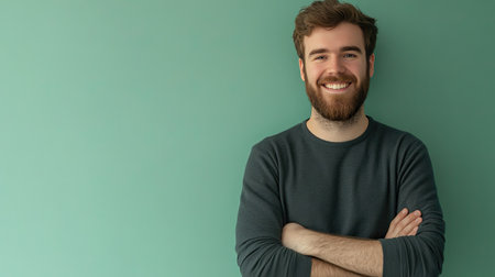 Smiling bearded young man with arms crossed, waist-up portrait against a pastel green background. Minimalist style with ample copy space.の素材