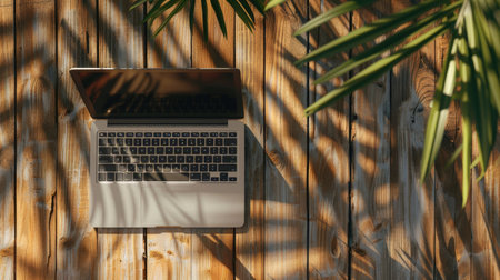 Top-down view of a laptop with a blank screen on a wooden background, highlighted by shadows of palm leaves. Perfect for summer digital concepts.の素材