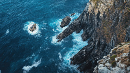 Lighthouse standing guard over a rocky coastline, with waves crashing in the blue ocean belowの素材
