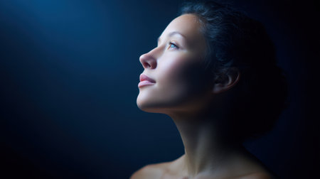 A peaceful image showcasing a young woman in profile, looking upwards as soft light highlights her serene expression against a dark backdrop, evoking introspection and beauty.の素材