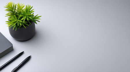 A modern workspace featuring a green plant, a notebook, and pens on a smooth grey surface, perfect for enhancing creativity and productivity in work or study environments.の素材