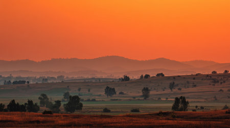 A breathtaking sunset casts vibrant orange and purple hues over rolling hills, showcasing layered mountains in the distance. The tranquil scene captures nature's serene beauty.の素材