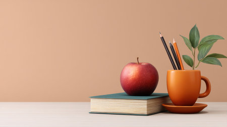 A vibrant apple rests on a stack of books next to an orange mug filled with pencils and featuring green leaves, perfect for educational themes.の素材
