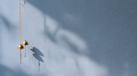 Two elegant gold bells hang gracefully against a light blue wall, casting delicate shadows and creating a serene atmosphere ideal for minimalist decor enthusiasts.の素材