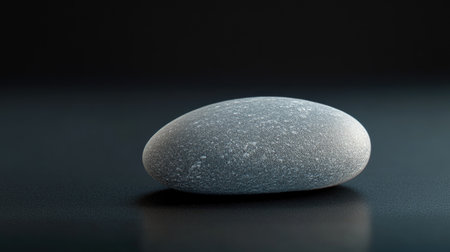 A smooth gray stone rests elegantly on a dark surface, showcasing its subtle texture and calm design. Ideal for themes of tranquility and nature.の素材