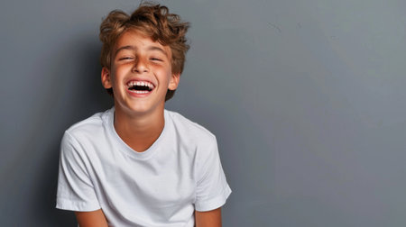 A cheerful young boy is captured laughing joyfully in a casual white t-shirt. His bright smile radiates happiness, set against a simple gray background.の素材