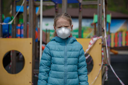 A little girl, wearing a medical mask on the background of the closed playground outdoor. A child in a protective mask. Warning of the dangers and safety quarantine measures against COVID-19. Coronavirus pandemic.の写真素材