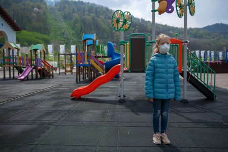 A little girl, wearing a medical mask on the background of the closed playground outdoor. A child in...